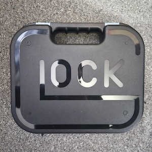 Puede incluir: Un maletín de transporte rígido y rectangular de color negro con un asa superior y esquinas redondeadas. La superficie frontal presenta la palabra LOCK en letras grandes de color negro brillante, con una línea horizontal.
