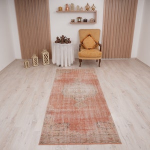 May include: A vintage, hand-woven rug in shades of peach, cream, and brown with a distressed, faded look. The rug has a rectangular shape and is laid out on a light wood floor.