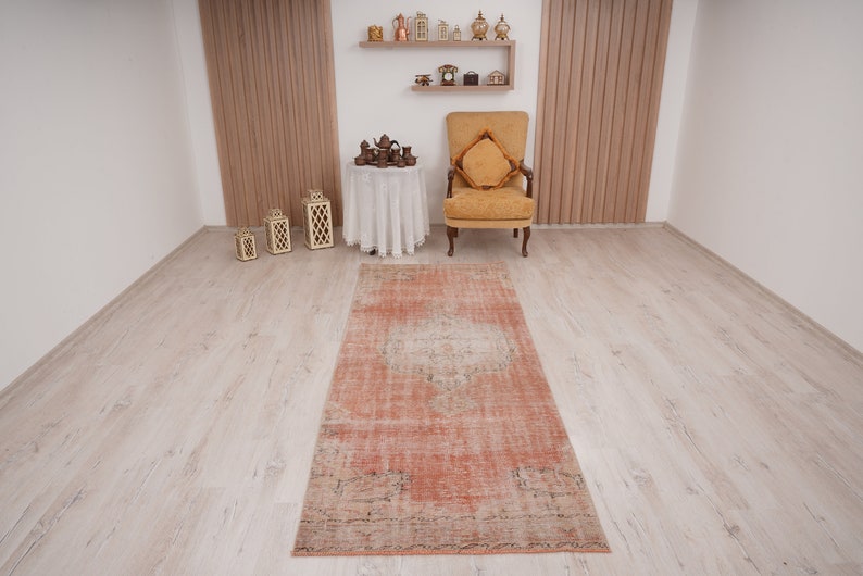 May include: A vintage, hand-woven rug in shades of peach, cream, and brown with a distressed, faded look. The rug has a rectangular shape and is laid out on a light wood floor.