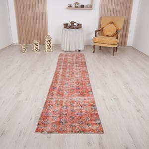 May include: A distressed, rectangular runner rug with a vintage appearance. The rug has a red and orange colour scheme with floral patterns and blue accents. It is placed on a light-coloured wooden floor. The rug's length is suitable for a hallway.