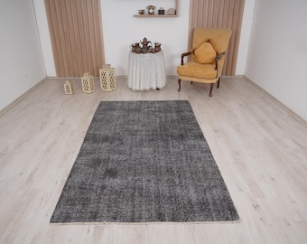 Puede incluir: Una alfombra rectangular gris oscuro con una apariencia desgastada y vintage. La alfombra está colocada sobre un suelo de madera de color claro. La habitación también cuenta con un sillón amarillo, una mesa pequeña y faroles decorativos.