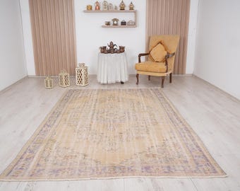 Può includere: Un tappeto vintage beige chiaro con un motivo floreale sbiadito e un bordo viola sottile. Il tappeto è steso su un pavimento in legno chiaro in una stanza con pareti bianche e una sedia.