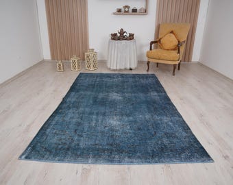 May include: A large, rectangular blue rug with a distressed, vintage appearance. The rug has a faded, mottled pattern with darker areas, creating a textured look. The rug is placed on a light-colored wooden floor.