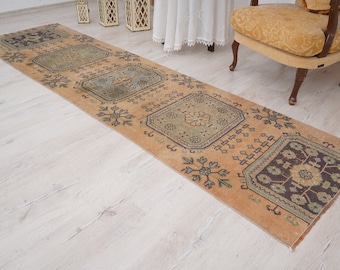 May include: A rectangular, vintage-style runner rug with a warm, earthy color palette. The rug features repeating geometric patterns in shades of brown, green, and blue. The rug is placed on a light-colored wooden floor.