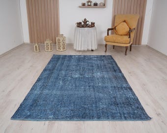 May include: A rectangular, distressed blue rug with a faded, vintage appearance. The rug is the focal point of the room, placed on a light-colored wooden floor. Decorative lanterns and a yellow armchair are also visible.