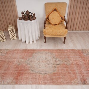 May include: A vintage, distressed, peach-colored rug with a faded, intricate design in the center. The rug is laid out on a light wood floor in a room with a white table and a yellow armchair.