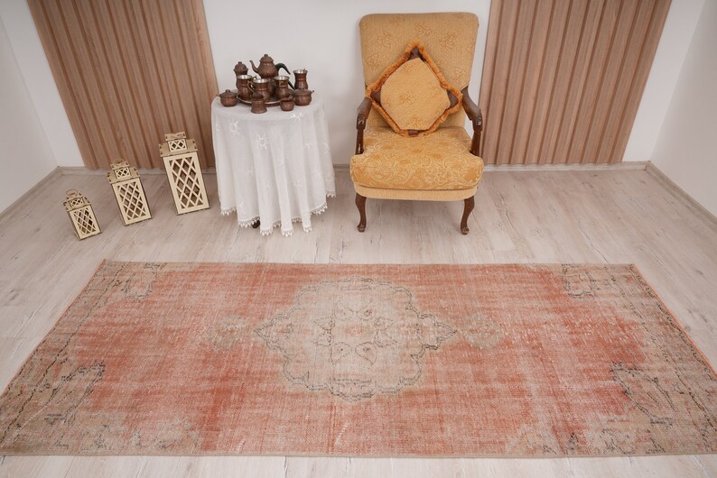 May include: A vintage, distressed, peach-colored rug with a faded, intricate design in the center. The rug is laid out on a light wood floor in a room with a white table and a yellow armchair.