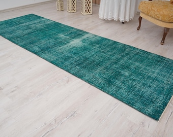 May include: A long, rectangular teal rug with a distressed, vintage appearance. The rug is placed on a light-colored wooden floor, with a glimpse of a chair and decorative lanterns in the background. The rug's dimensions are not visible.