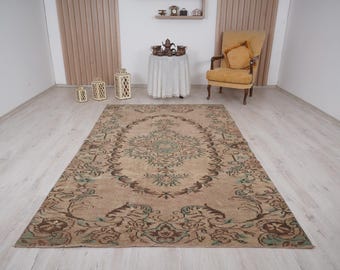 May include: A rectangular, vintage-style rug with an ornate, floral pattern in shades of brown, beige, and teal. The rug is placed on a light-colored wooden floor, with a chair and side table in the background. The rug's design features a central medallion.
