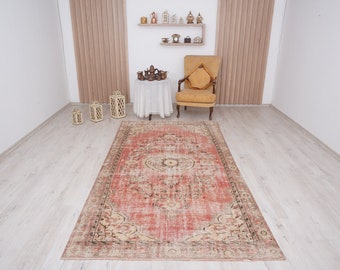 May include: A vintage-style rug with a faded red background and intricate floral patterns in beige and green. The rug is laid out on a light wood floor in a room with a chair and decorative items.