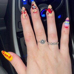 May include: A hand with long, almond-shaped nails painted with different designs. The nails are painted with red, white, blue, yellow, and black. The designs include a heart, an eye, and the word "Good".