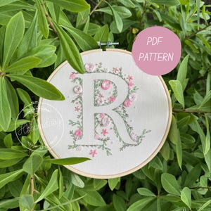 May include: A floral embroidery hoop featuring the letter "R" adorned with pink flowers and green leaves. The design includes small, decorative buttons. The text "PDF PATTERN" is in a pink circle. The background is green foliage.