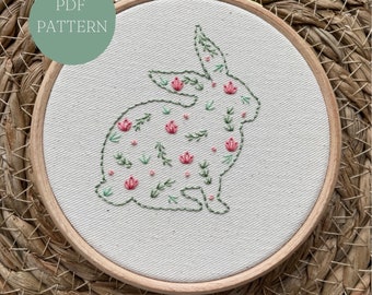 May include: A white embroidery hoop with a floral design of a rabbit. The rabbit is outlined in green thread and filled with pink and green flowers.