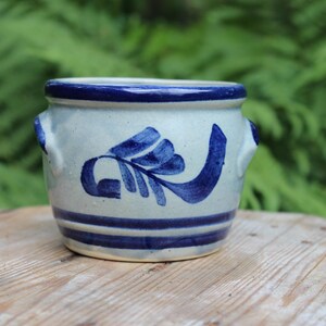 May include: A small, white ceramic pot with blue painted designs. The pot has two small handles on the sides and a blue rim.