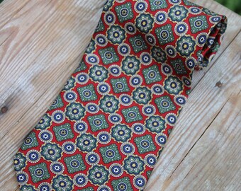 May include: A red silk tie with a green, blue, and gold patterned design. The pattern features repeating geometric shapes and floral motifs.