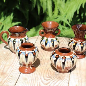 Può includere: Cinque piccoli vasi in ceramica marrone con intricati disegni dipinti. I vasi hanno forme e dimensioni diverse e sono tutti decorati con un motivo simile di linee e punti bianchi, blu e gialli.