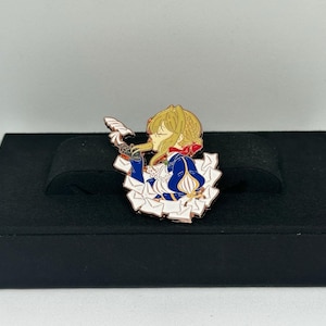 May include: A colorful enamel pin featuring a character with blonde hair, wearing a blue and white dress, holding a feather. The pin has gold-colored trim and is set against a black display box. The character is from an anime.