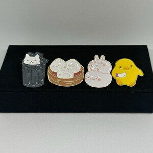 Könnte beinhalten: Vier Emaille-Pins auf schwarzem Hintergrund. Die Pins zeigen eine Katze in einem Mülleimer, einen Korb mit gedämpften Brötchen, ein Kaninchen mit Schleife und eine gelbe Ente, die ein Messer hält.