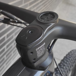 May include: Close-up of a black bicycle handlebar with a black and silver stem. The stem has a small black device mounted on top and the text "CP0005" is printed on the side.