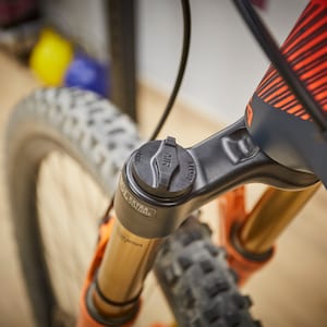 Può includere: Primo piano di una forcella per mountain bike nera con una decalcomania arancione e nera. La forcella ha un tappo nero per la valvola dell'aria con la scritta "AIR" stampata su di esso.