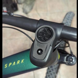 May include: Close-up of a black bicycle handlebar with a black and gray stem. The stem has a round, black mount for a cycling computer. The handlebar is labeled "SPARK" in yellow lettering on a green background.