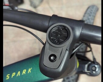 May include: Close-up of a black bicycle handlebar with a black and grey stem. The stem has a round, black mount for a cycling computer. The handlebar is labelled "SPARK" in yellow lettering on a green background.