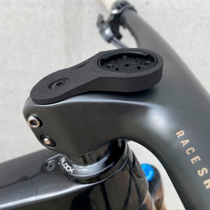 Peut inclure: Gros plan sur une potence de vélo noire avec un support d'ordinateur noir et gris. La potence a une vis argentée et le texte "RACE SHOP" est visible sur le cadre du vélo.