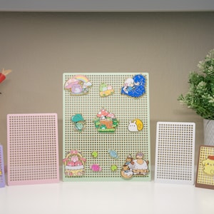 May include: A collection of colourful enamel pins displayed on green, pink and white pegboards. The pins feature cartoon characters and designs. The pegboards are arranged on a white surface with a small sign that reads "From a Cat".