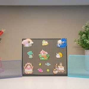 Puede incluir: Una rejilla negra con pines de esmalte de colores con personajes de dibujos animados y diseños. Los pines están dispuestos en un patrón de rejilla sobre un fondo negro. También hay dos rejillas más pequeñas, gris y azul, en la mesa. Un letrero dice "From a Cat".