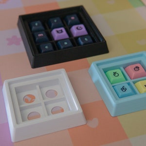 May include: Three different keycap trays, one black, one white, and one light blue. The trays are empty except for the light blue tray, which has four keycaps with fruit designs. The keycaps are green, pink, light blue, and yellow.
