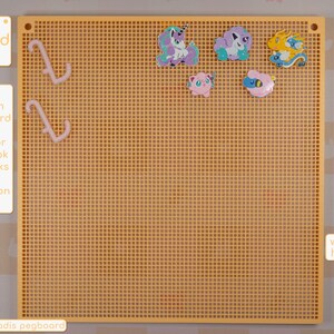 以下が含まれることがあります： 黄色いメガペグボード、幅305mm、高さ305mm、格子模様。ピンク色のフック2本と、カラフルな漫画のキャラクターの装飾がいくつか含まれています。ボードにはサイズとフックの色オプションが表示されています。