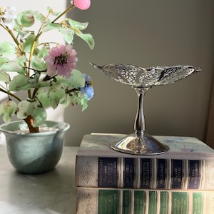 May include: A silver-toned pedestal bowl with a delicate, openwork filigree rim, resting on a stack of two vintage books. The top book spine reads "Reader's Digest Condensed Books" and the bottom book spine also reads "Reader's Digest Condensed Books".