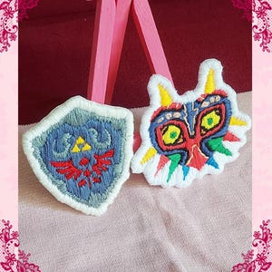 Peut inclure: Deux écussons brodés avec des motifs de jeux vidéo. L'un est un bouclier bleu avec un emblème rouge et des détails jaunes. L'autre est un masque coloré avec des détails jaunes, rouges, bleus et verts, soulignés de blanc.