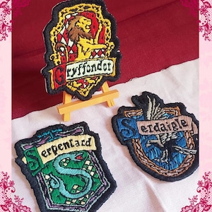 Peut inclure: Écussons brodés représentant les blasons de Gryffondor, Serpentard et Serdaigle. L'écusson de Gryffondor montre un lion, celui de Serpentard un serpent et celui de Serdaigle un aigle. Chaque écusson a une bordure noire et est sur un fond rouge et blanc.