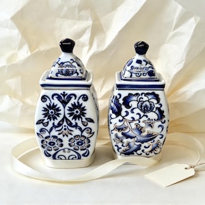 May include: Two square porcelain jars with lids, featuring intricate blue and white floral patterns with gold accents. Each jar has a tapered top and a smooth, glossy finish, standing side by side as a matching set of decorative containers.