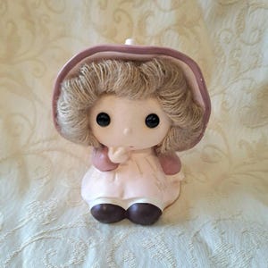 May include: A ceramic figurine of a girl with brown hair wearing a pink dress and a pink bonnet. She is holding her hand to her chin and has a sweet expression on her face.