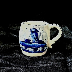 May include: A small white ceramic mug with a blue and white windmill design. The mug has a handle and a rounded shape.