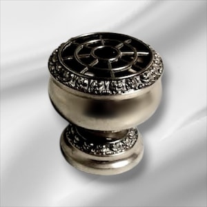 May include: Silver-toned metal flower vase with an ornate design and a circular wire mesh insert. The vase has a round base and a wide, bowl-shaped top.