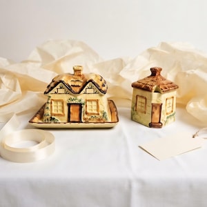 May include: A ceramic butter dish and a matching lidded jar, both shaped like small cottages with thatched roofs. The pieces feature cream-coloured walls, brown doors, and painted green foliage details, set against a neutral background.