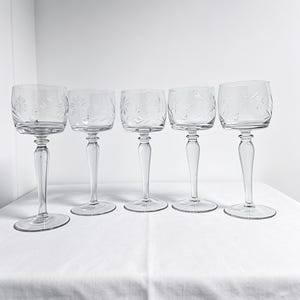 May include: A set of five clear glass stemmed goblets, each featuring a delicate etched starburst or floral pattern around the upper bowl. The glasses have a smooth, rounded rim, a slender stem with a slight bulbous detail, and a circular base.