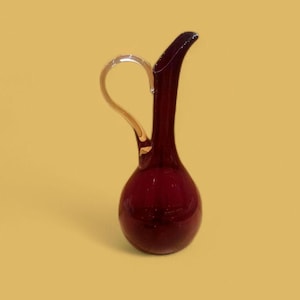 May include: A deep red glass jug with a gold-coloured handle. The jug has a slender neck and a rounded base. The handle curves from the top to the side. The jug is set against a yellow background.