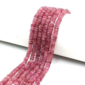 May include: Strands of pink tourmaline beads. The beads are small, square-cut, and have a consistent color throughout. The beads are strung together, creating a flowing, textured appearance. The background is white.