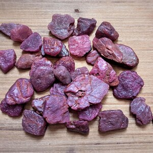 Könnte beinhalten: Eine Ansammlung von rohen, ungeschliffenen Rubin-Edelsteinen in verschiedenen Schattierungen von tiefem Magenta, Dunkelrosa und Violett. Diese natürlichen Mineralsteine weisen unregelmäßige, gezackte Formen mit texturierten, matten Oberflächen auf.