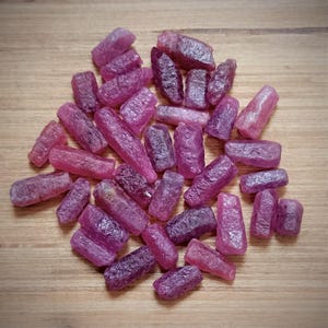 Könnte beinhalten: Eine Ansammlung von rohen, ungeschliffenen Rubinkristallen in verschiedenen Rosa- und Dunkelrottönen. Diese natürlichen Edelsteine weisen unregelmäßige, längliche und blockartige Formen mit texturierten, matten Oberflächen auf, locker verstreut.