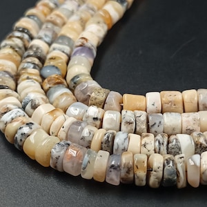 May include: A strand of small, disc-shaped beads made of natural stone in shades of cream, beige, tan, and soft gray. The beads feature organic, speckled patterns and translucent inclusions, arranged in a curved line on a dark surface.