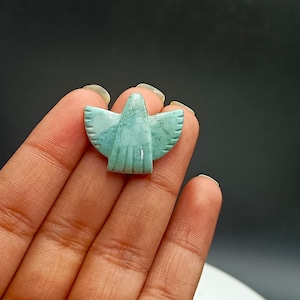 May include: A small, turquoise-coloured carved stone in the shape of a bird with outstretched wings. The bird has detailed feather carvings and is held in a hand against a dark background. The stone has a smooth, polished finish.