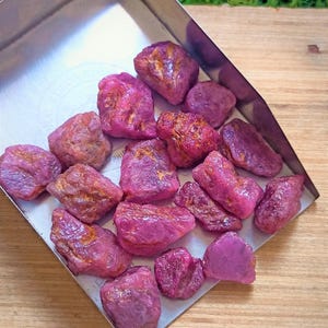 May include: A collection of raw, unpolished ruby gemstones with a deep pinkish-red hue and irregular, textured surfaces. These small, natural stones are contained within a silver-colored metal tray, displaying varied shapes and sizes.