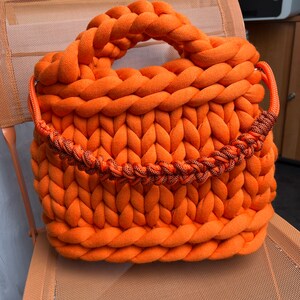 Puede incluir: Un bolso tote de punto grande de color naranja brillante con un asa gruesa de cuerda. El bolso está hecho de hilo grueso y tiene una textura tejida.