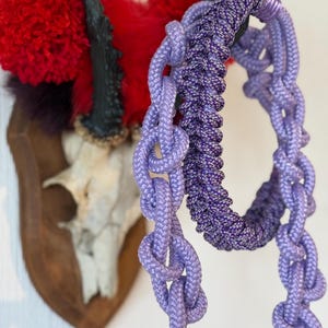 Könnte beinhalten: Nahaufnahme von zwei lila Seilartikeln, einer in Kettenform und der andere ein geflochtener Kreis. Im Hintergrund ein Hirschschädel auf einer Holzplatte, verziert mit roten und violetten Akzenten.