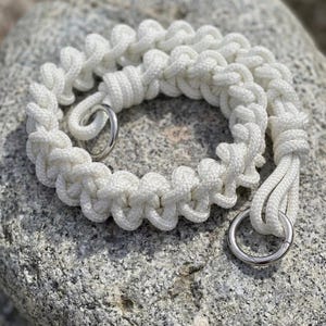 Può includere: Un braccialetto in paracord bianco caratterizzato da un complesso motivo a nodi intrecciati e due anelli in metallo color argento lucido. La corda è intrecciata in una spessa fascia circolare testurizzata con nodi sicuri ai punti di giunzione.